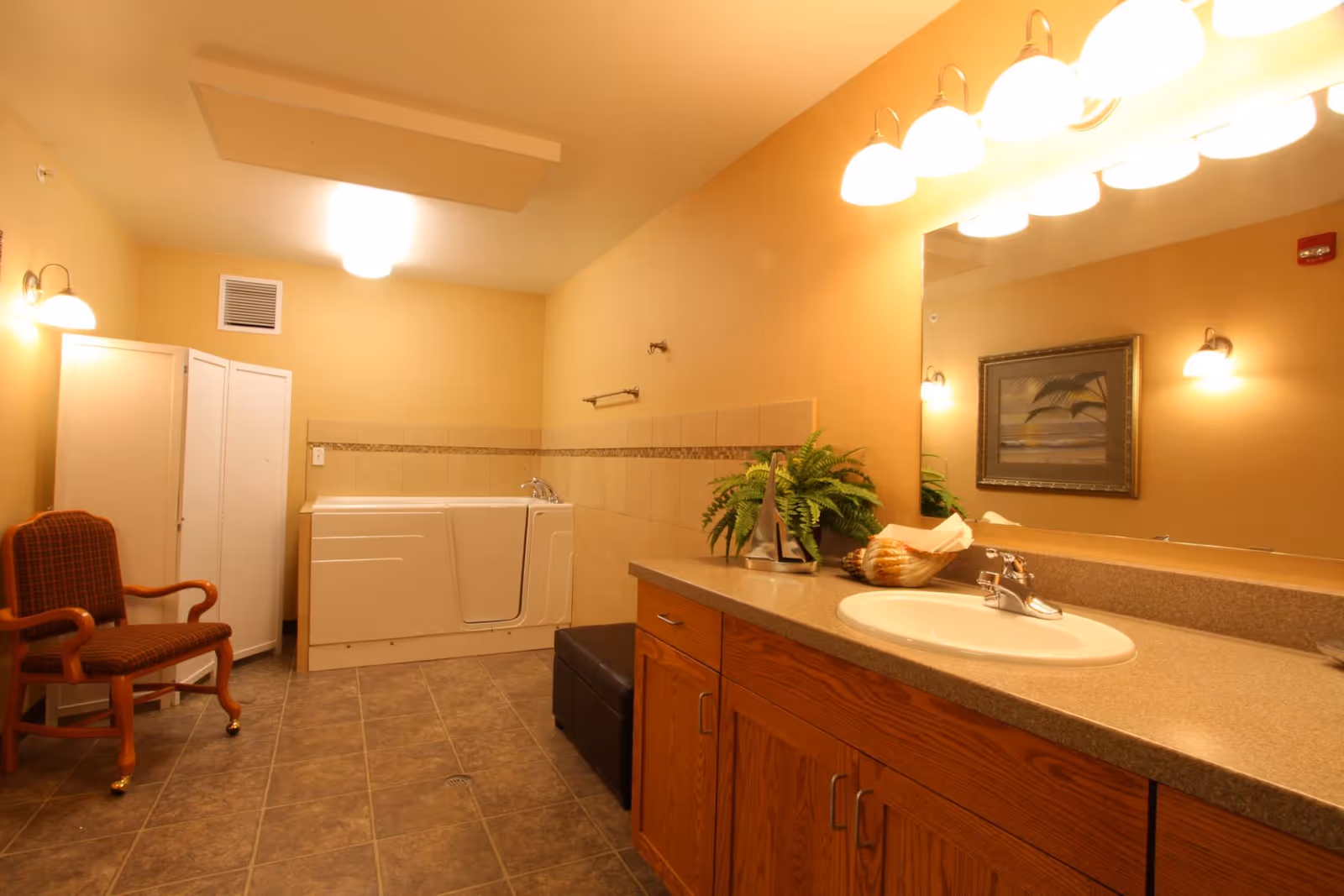 Bright, accessible bathroom featuring a walk-in tub, vanity with sink and large mirror, wooden cabinets, and a chair.