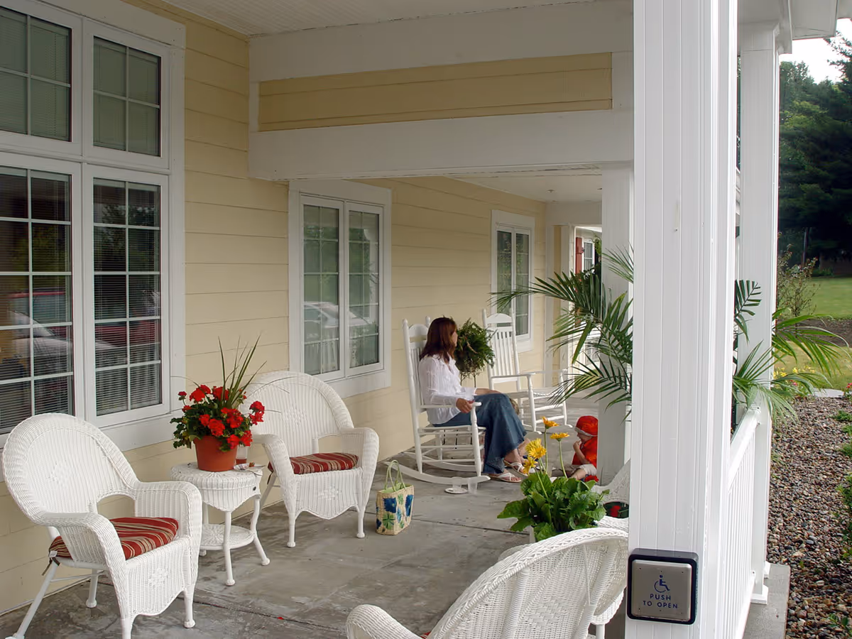 A covered porch area with white wicker chairs and a small table with a potted plant. A woman is sitting on a white rocking chair, and a child is nearby. There are several plants and flowers decorating the porch, and a push button for automatic door opening is mounted on a white column.