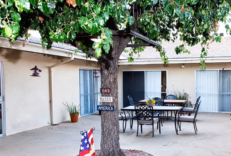 Outdoor courtyard area with a large tree in the center displaying a sign that reads 'God Bless America'. Surrounding the tree are several metal tables and chairs with cushions. The courtyard is enclosed by a beige building with sliding glass doors and potted plants.