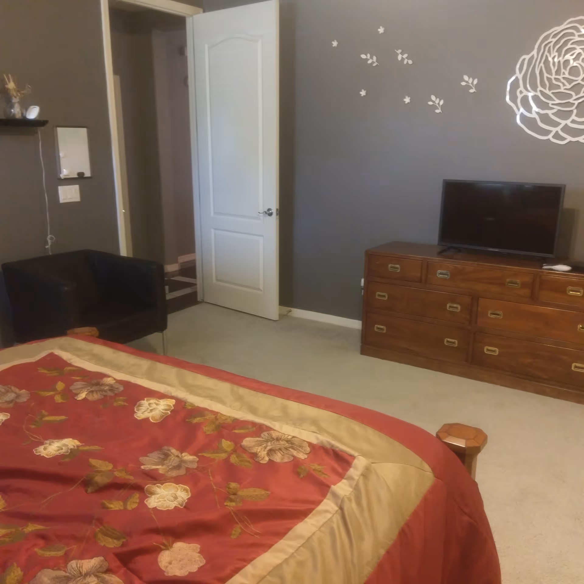 Bedroom with a floral bedspread, wooden dresser and TV, a black armchair, and an open door against gray walls.