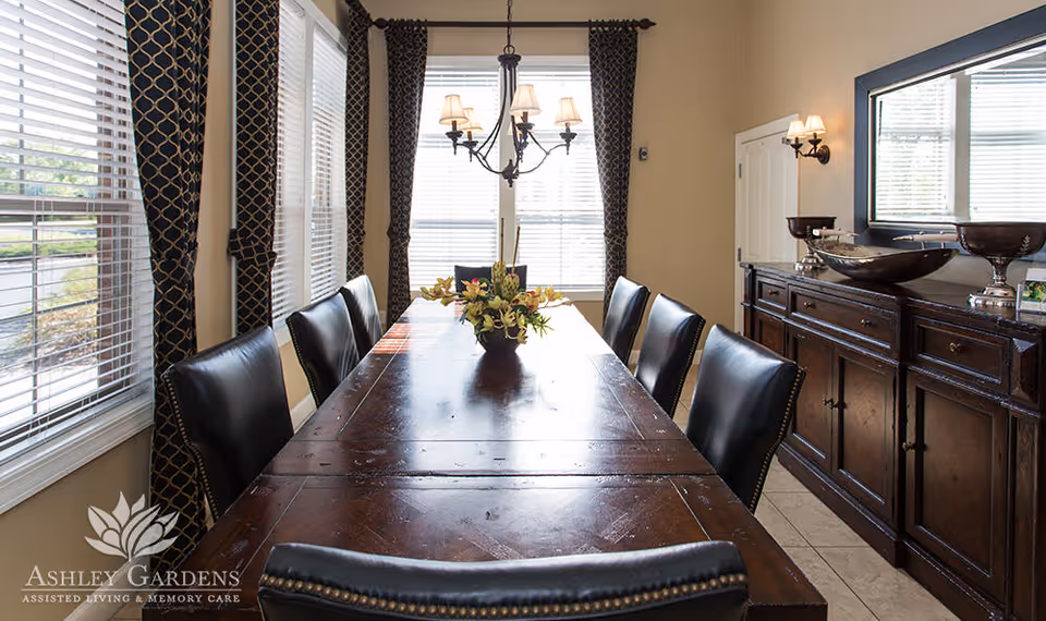 A dining room with a long wooden table surrounded by eight black leather chairs. The table has a floral centerpiece. The room features large windows with patterned curtains, a chandelier hanging above the table, and a dark wooden sideboard with decorative bowls and a large mirror above it.