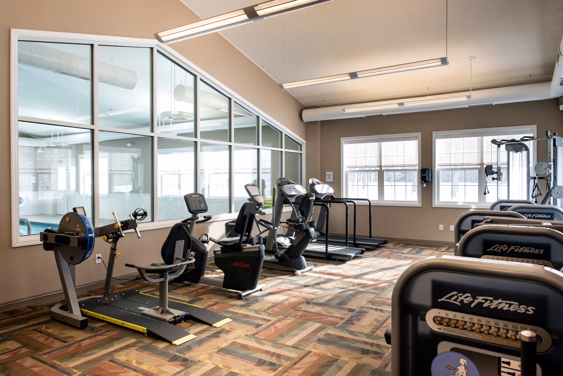 A fitness room with various exercise equipment including stationary bikes, treadmills, and weight machines. The room has large windows letting in natural light and a glass wall looking into an adjacent indoor pool area. The floor is covered with patterned carpet and the walls are painted beige.