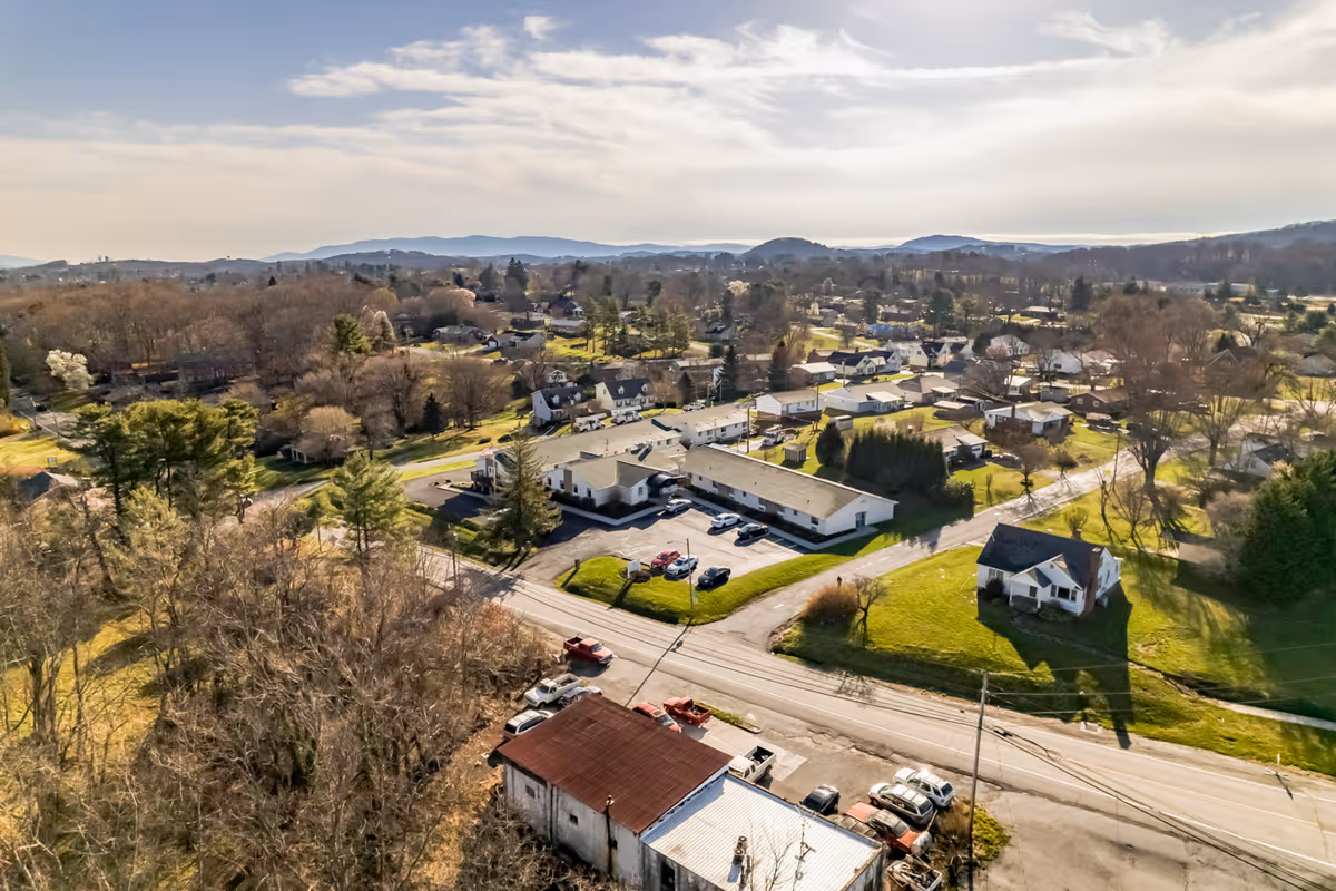 Aerial view of a suburban area with houses, trees, and roads under a partly cloudy sky. The landscape includes a mix of residential buildings, some with large yards, and a few parked cars along the streets. Hills and mountains are visible in the distance.