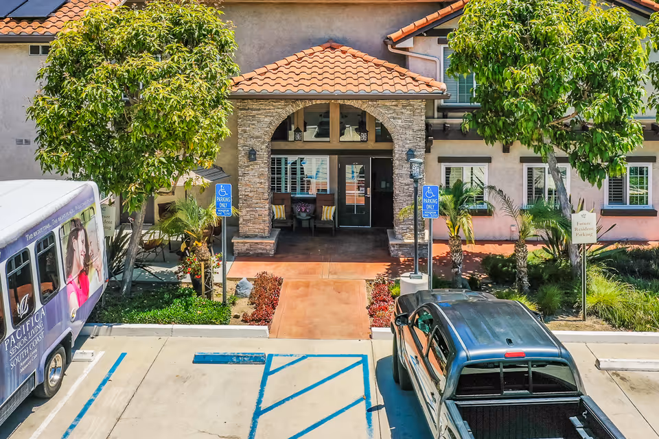 Entrance of Pacifica Senior Living South Coast showing a tiled-roof porch with a stone arch, accessible parking spaces, a shuttle van, and a pickup truck.