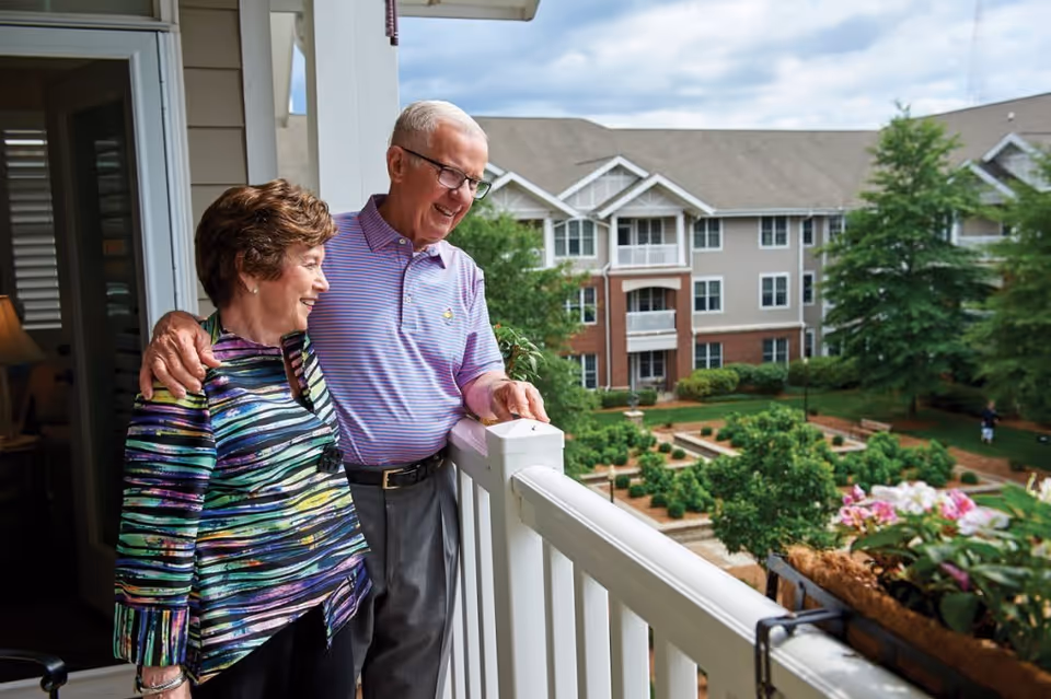 An elderly couple stands on a balcony overlooking a landscaped garden with shrubs and flowers. The man has his arm around the woman, and both are smiling while enjoying the view of the residential building and greenery in the background.