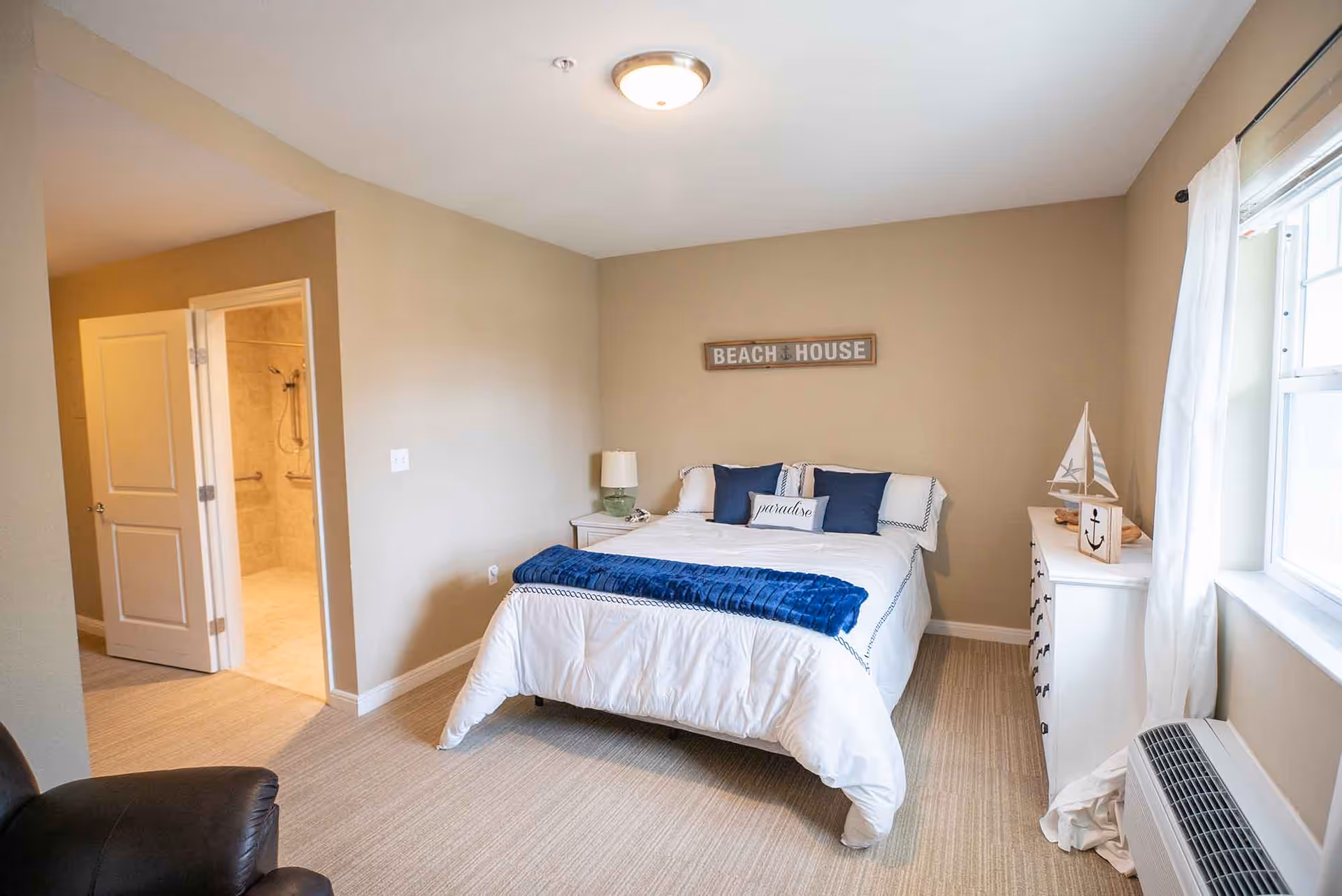 A cozy bedroom with a neatly made bed featuring white bedding and blue accent pillows and throw. Above the bed is a wall sign that reads 'BEACH HOUSE'. To the right of the bed is a white dresser with nautical decor including a small sailboat and an anchor block. A window with white curtains allows natural light into the room. To the left, an open door reveals a bathroom with tiled walls and a shower. The room has beige walls and carpeted flooring.