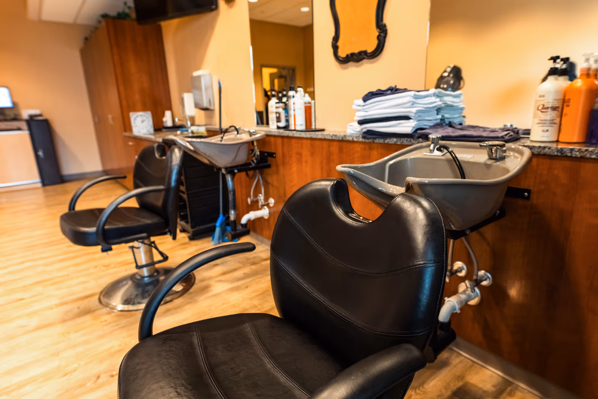 Interior view of a hair salon area in a senior living facility with two black salon chairs in front of wash basins, a countertop with folded towels and hair care products, and a large mirror on the wall.