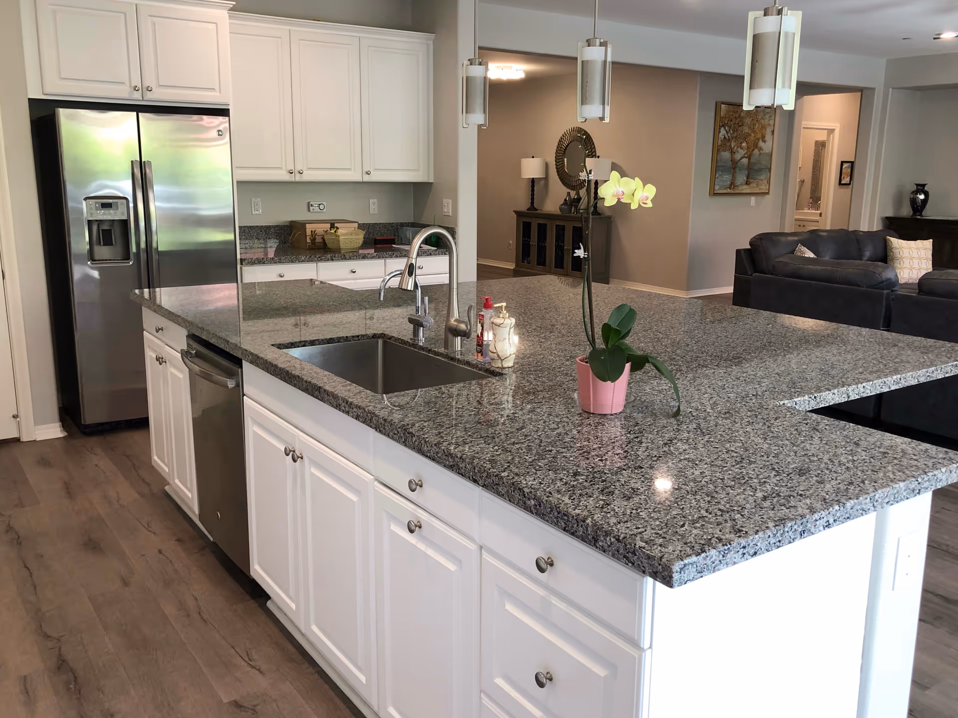 Modern kitchen with white cabinets, granite countertops, a stainless steel refrigerator, dishwasher, and a sink on a large island. A pink orchid plant is placed on the island. In the background, there is a living room area with dark sofas, a lamp, and wall art.