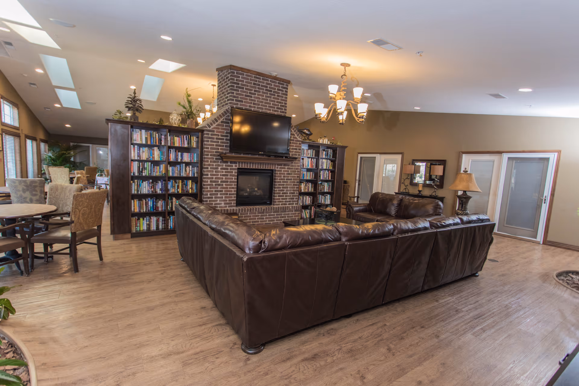 A spacious living room area with a large brown leather sectional sofa facing a brick fireplace with a mounted flat-screen TV above it. There are bookshelves filled with books on either side of the fireplace. The room has wooden flooring, beige walls, and several windows allowing natural light. A chandelier hangs from the ceiling, and there are additional seating areas with tables and chairs visible in the background.