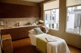 A well-lit treatment room with a massage or therapy bed covered with a white quilt and a green throw blanket. There is a patterned pillow on the bed and a folded towel at the foot. The room has wooden cabinets with a mosaic tile backsplash, a wicker basket, and large windows letting in natural light with a view of an adjacent building.