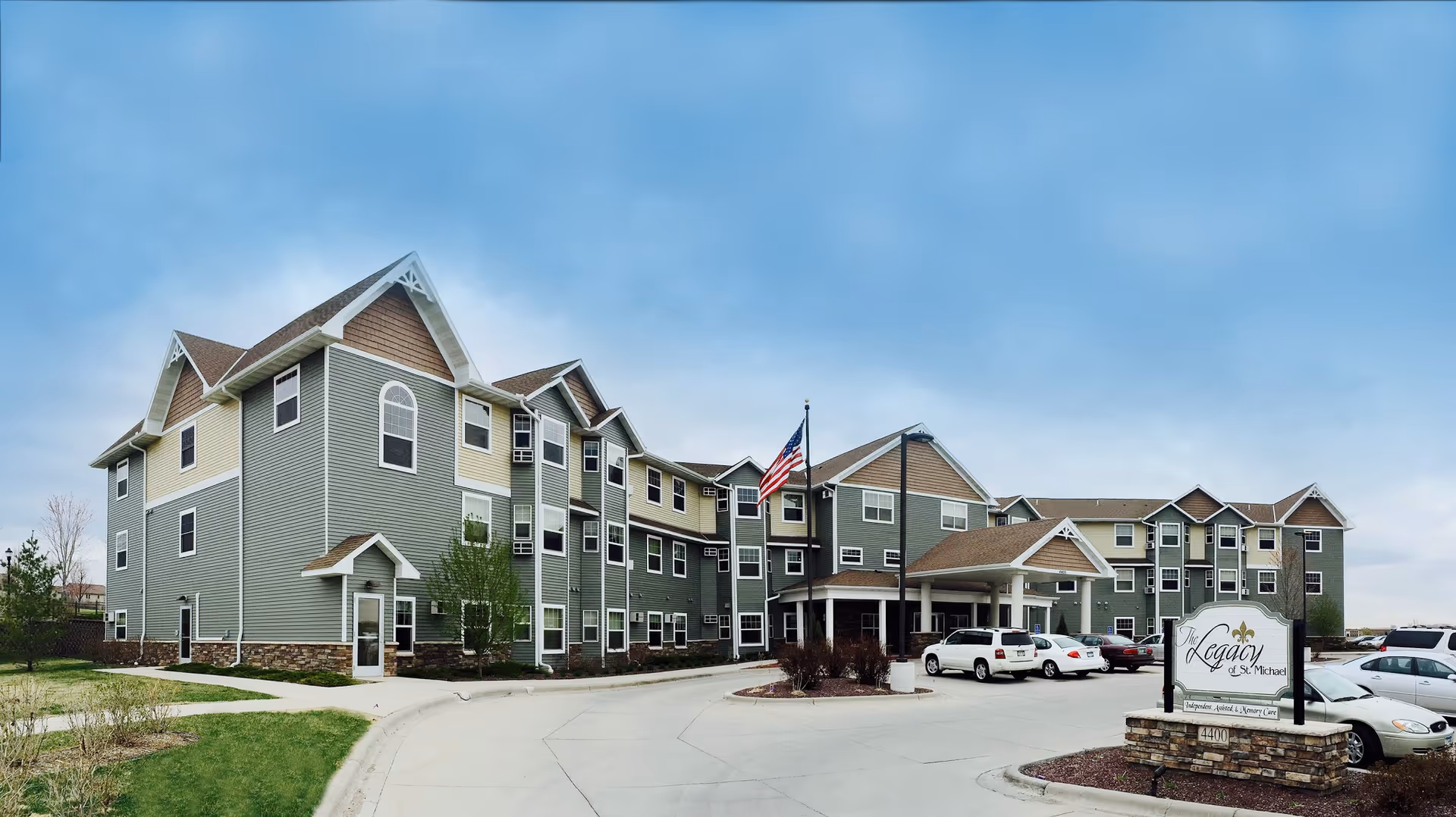 Exterior view of a three-story senior living facility building with gray and beige siding, multiple windows, and a covered entrance. Several cars are parked in front, and an American flag is flying near the entrance. A sign in the foreground reads 'Legacy of St. Michael'.