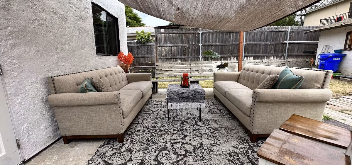 Outdoor patio area with two beige tufted sofas facing each other on a patterned rug, a small table with a decorative cloth and candle in the center, a wooden bench in the background, and a shade cloth overhead. The area is enclosed by a wooden fence and adjacent to a white building wall with a window.