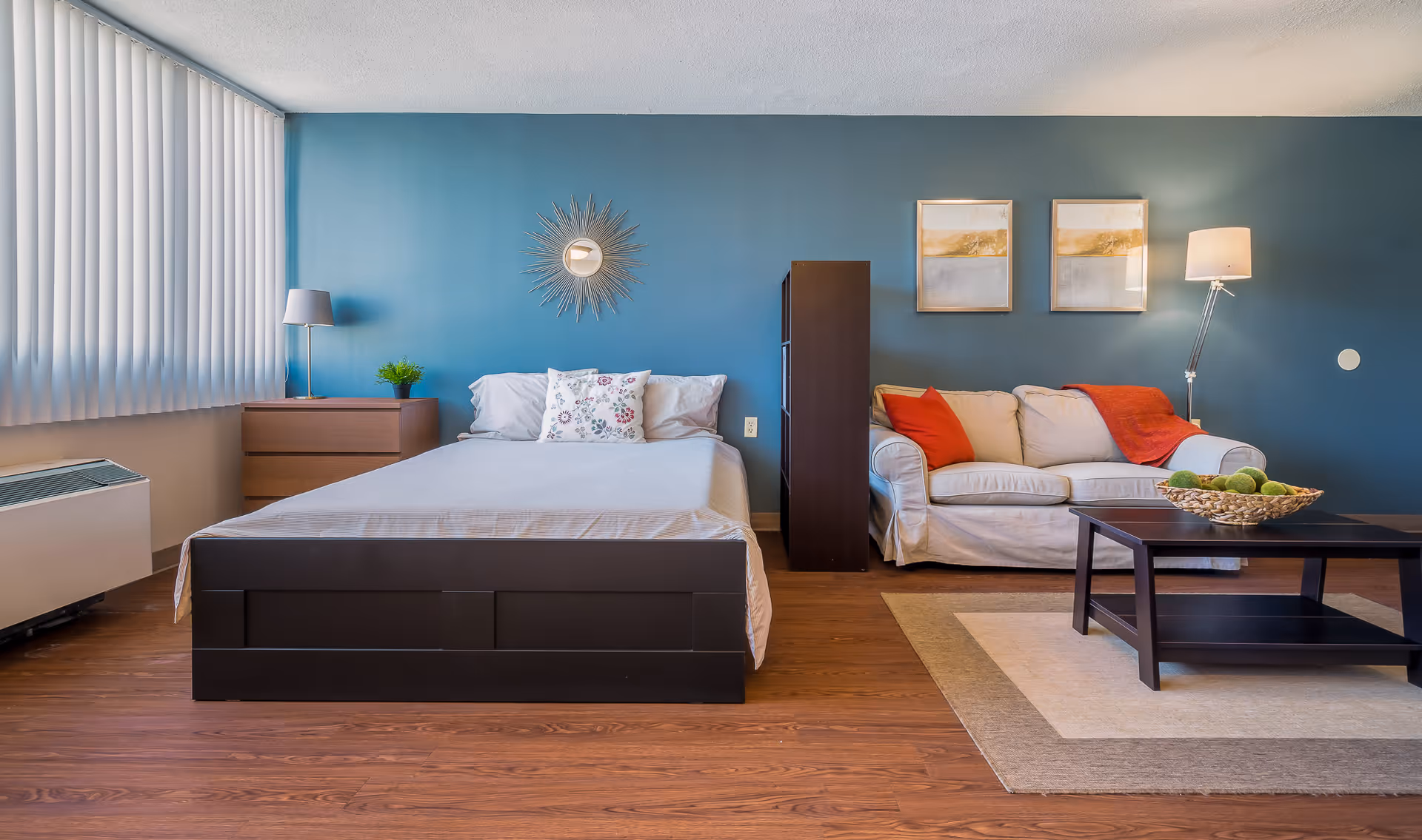 A cozy studio apartment with a bed on the left side featuring white bedding and a floral pillow. Next to the bed is a wooden nightstand with a lamp and a small plant. On the right side, there is a white sofa with red cushions and a red throw blanket. A dark wooden coffee table with a decorative bowl of green apples sits on a beige and cream area rug. The walls are painted blue with two framed pictures and a sunburst mirror above the bed. The floor is wooden, and vertical blinds cover the window on the left.
