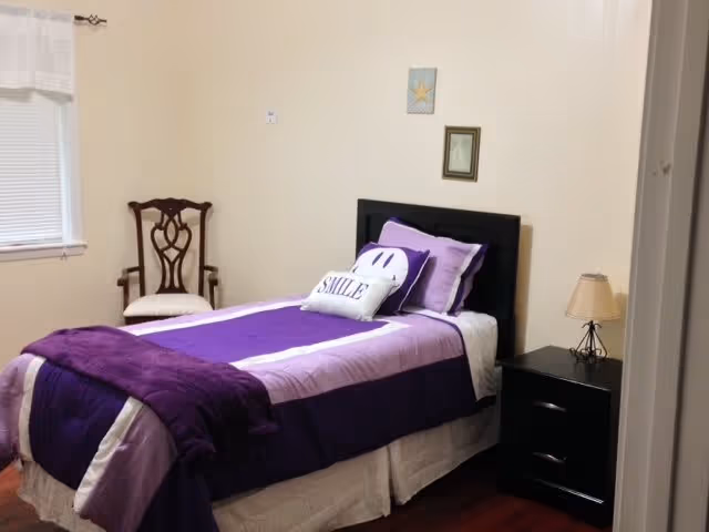 A small bedroom with a single bed dressed in purple and white bedding, including a pillow with the word 'SMILE' and a smiley face pillow. There is a wooden chair with a cushioned seat near the window with closed blinds. A black nightstand with two drawers and a beige lamp sits next to the bed. Two small framed pictures hang on the wall above the headboard.