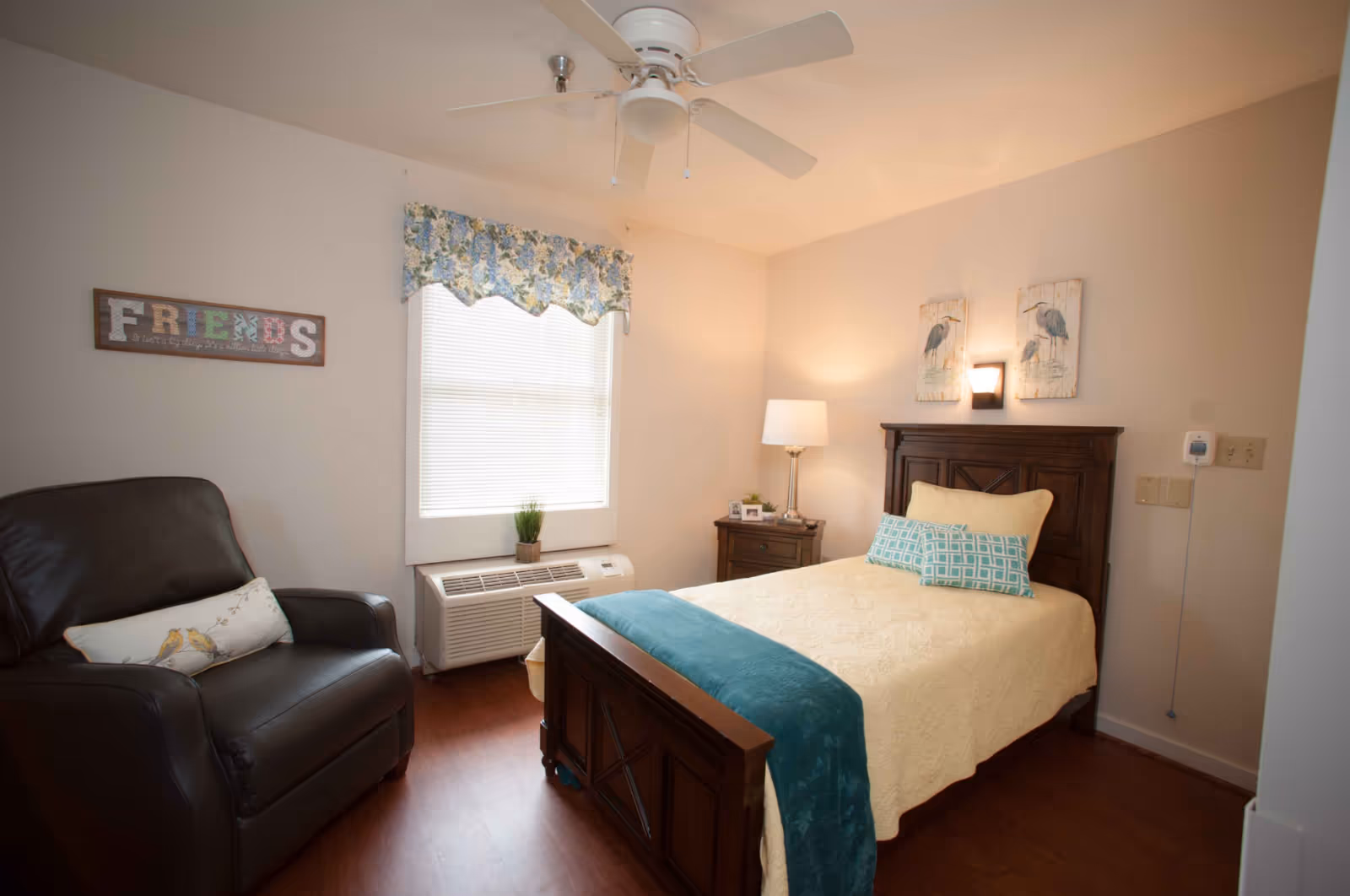 A cozy bedroom in a nursing home featuring a wooden bed with a cream-colored quilt and teal throw blanket, two decorative pillows, a wooden nightstand with a lamp and small framed photos, a leather armchair with a decorative pillow, a window with floral valance, and wall art including a 'FRIENDS' sign and two bird paintings.