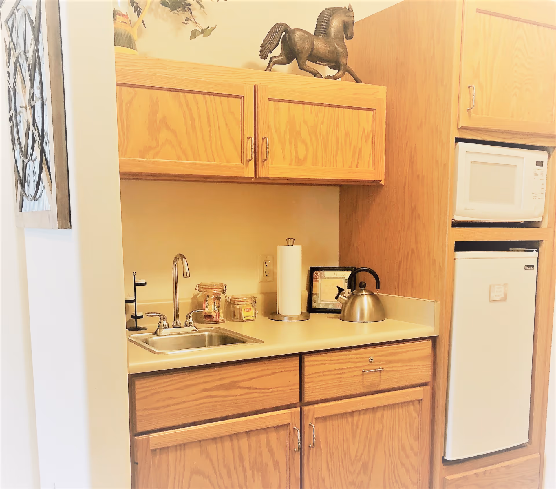 A small kitchenette with wooden cabinets, a stainless steel sink with a faucet, a paper towel holder, a metal kettle, two glass jars, and a microwave above a compact refrigerator. A decorative horse statue is placed on top of the upper cabinets.