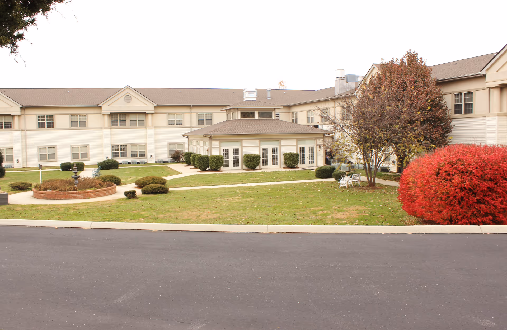 Exterior view of Cedar Hills Senior Living facility showing a two-story building with multiple windows, a central pavilion with glass doors, landscaped green lawn, bushes, trees with autumn foliage, and a paved driveway in the foreground.