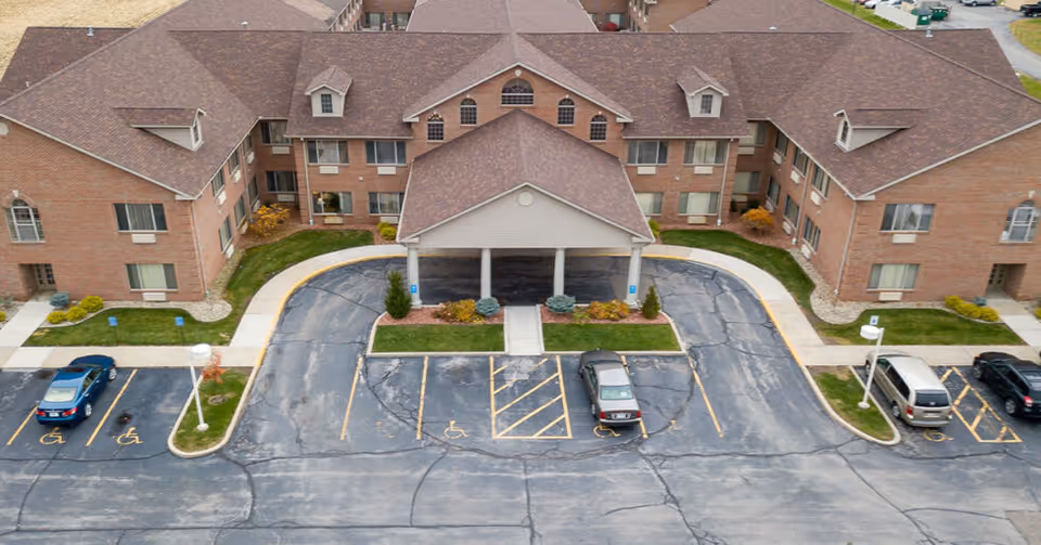 Aerial view of the front entrance of a large brick senior living facility with a covered drop-off area, multiple windows, and a parking lot with several cars and designated handicapped parking spaces.