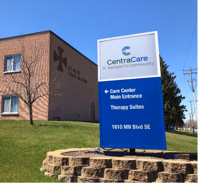 Outdoor view of a brick building with two windows and a leafless tree on a grassy lawn. In front of the building is a large blue and white sign for CentraCare St. Benedict's Community indicating the Care Center Main Entrance and Therapy Suites at 1810 MN Blvd SE. The building has a cross symbol and the words 'LET ALL BE TREATED AS CHRIST' on the wall.