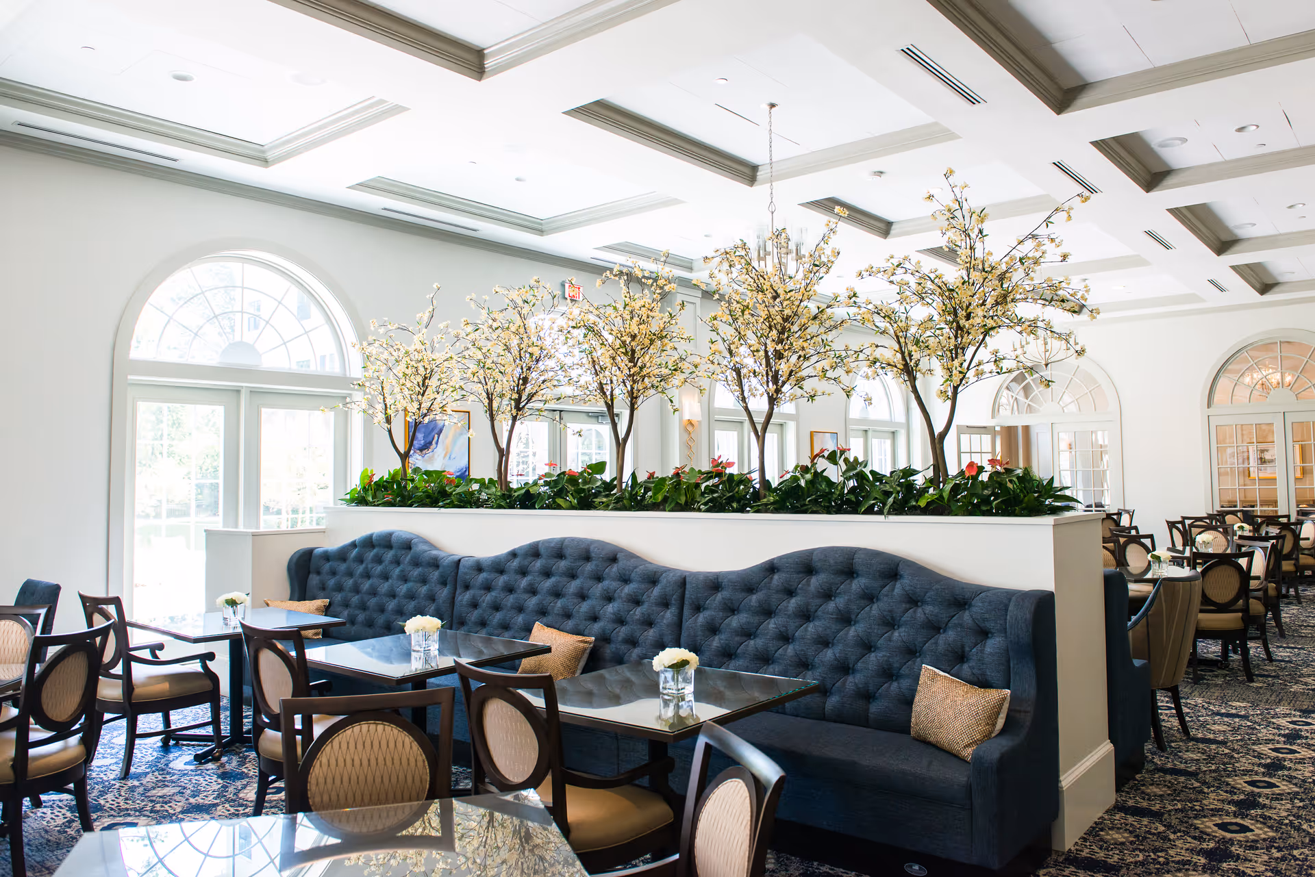 Bright and elegant dining room with tufted blue banquette seating, dark wood chairs with beige cushions, glass-top tables, and decorative artificial trees with white blossoms in planters dividing the space. Large arched windows allow natural light to fill the room.