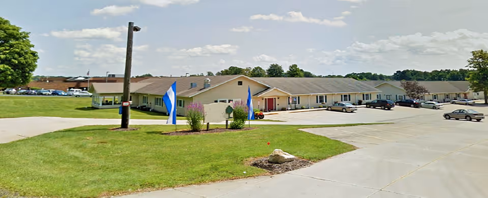 Exterior view of a single-story building with a parking lot in front, green lawn, and a few flags near the entrance under a partly cloudy sky.