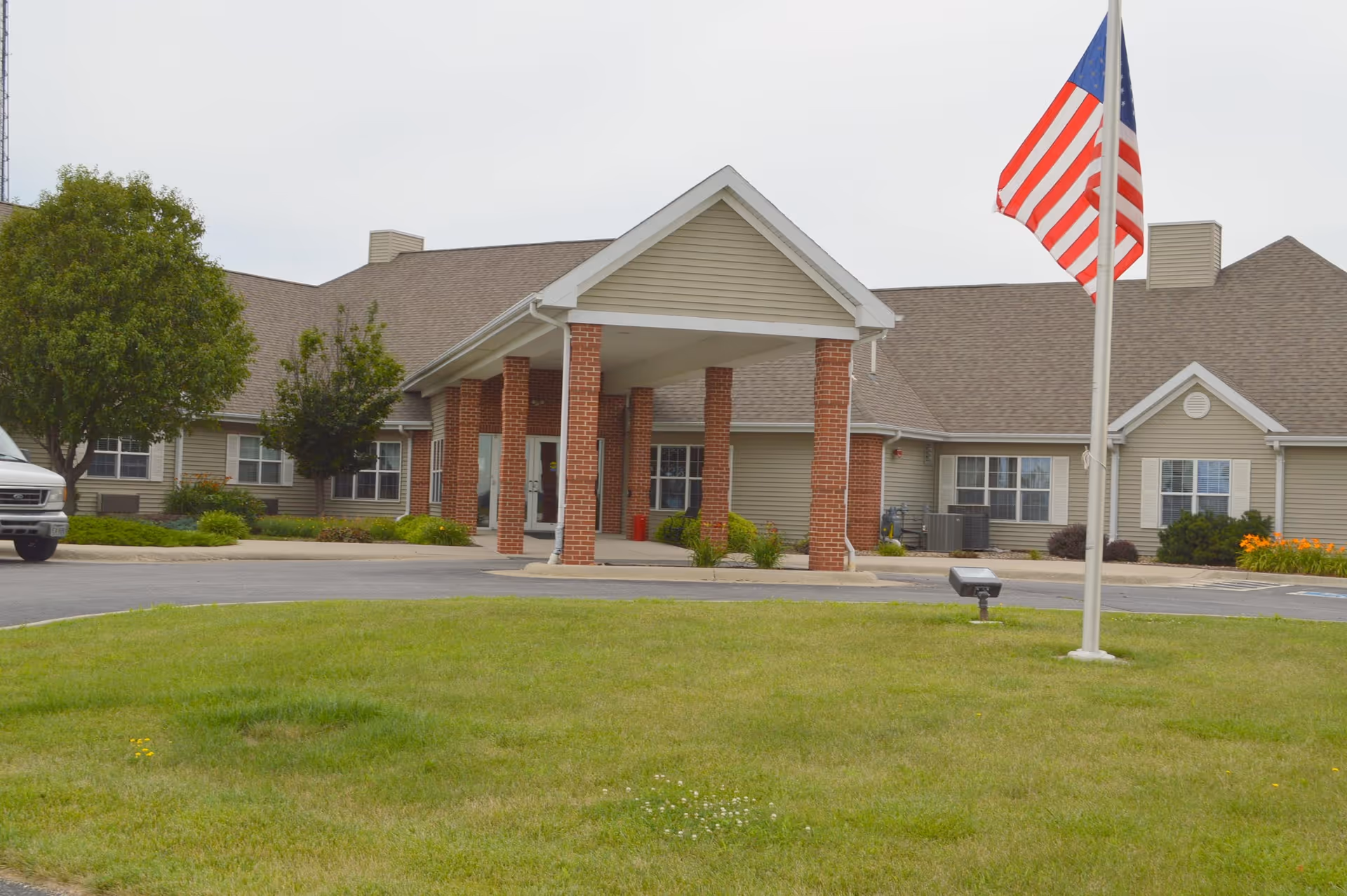 Front exterior view of Avoca Lodge, a single-story building with beige siding and a brown roof. The entrance features a covered portico supported by brick columns. There is a green lawn in the foreground and an American flag on a flagpole to the right. Trees and shrubs are planted around the building.