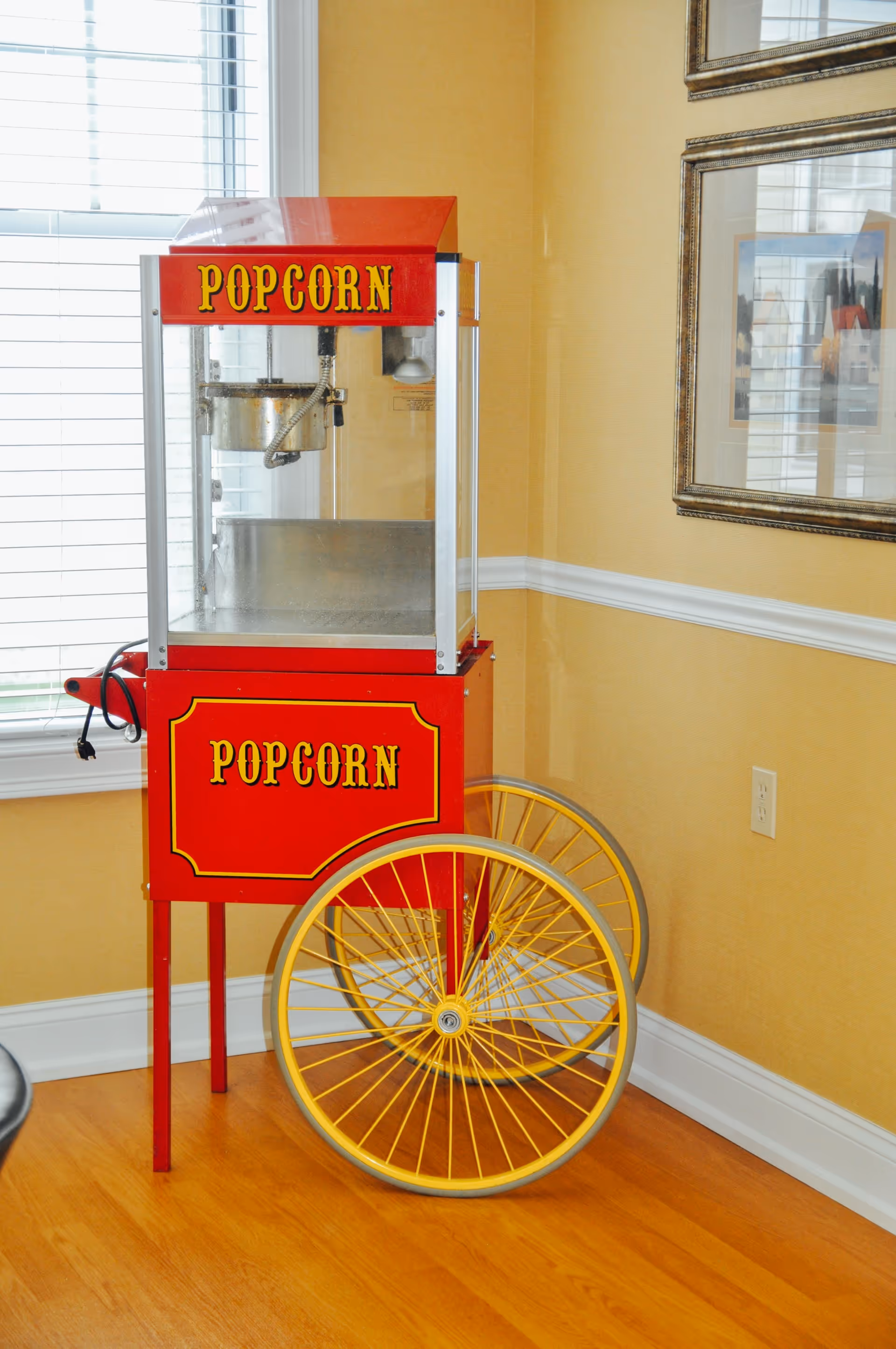 A vintage-style red popcorn machine with yellow wheels is placed in the corner of a room with wooden flooring and yellow walls. There is a window with white blinds on the left and framed artwork on the wall to the right.