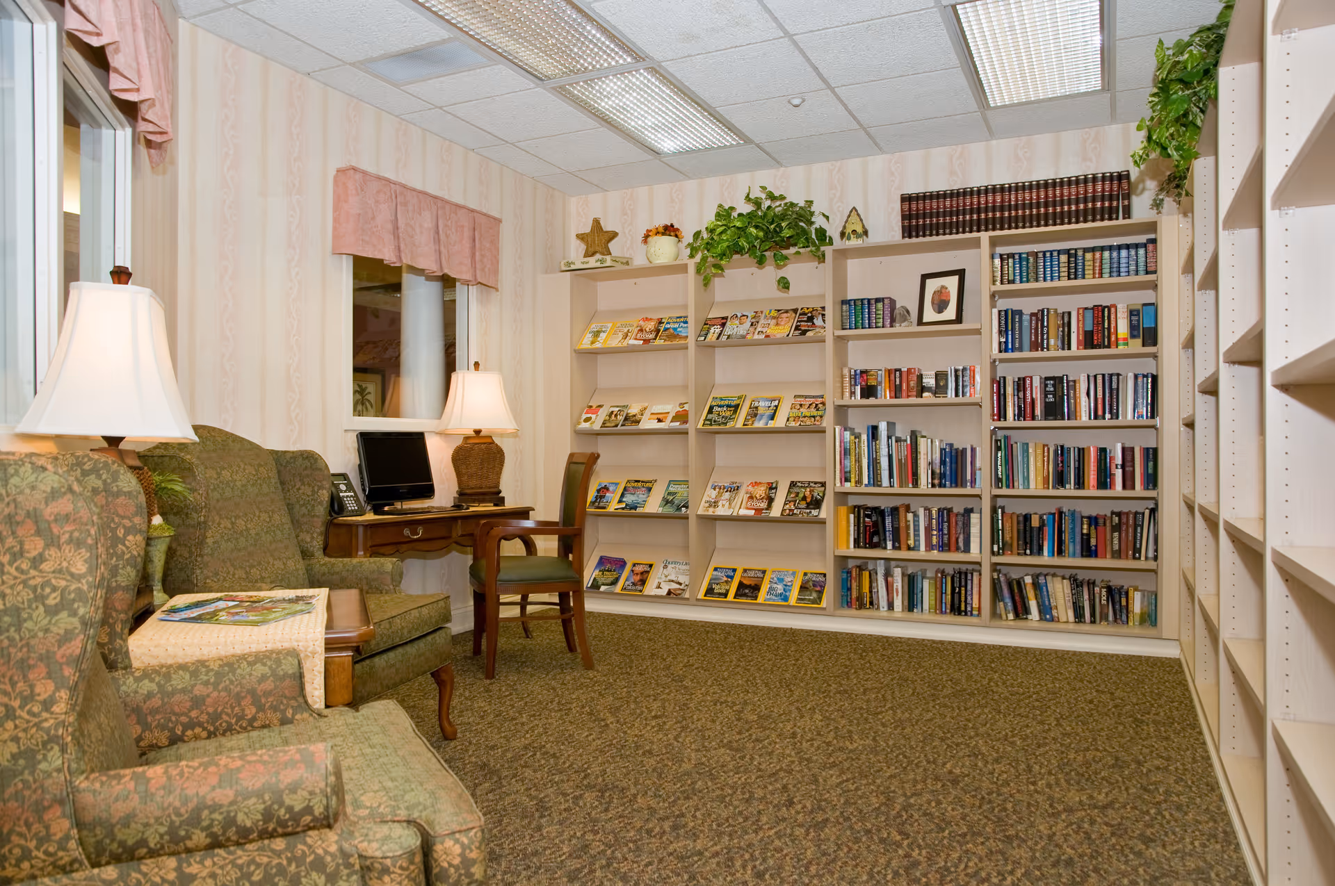 Cozy reading room with armchairs, table lamps, a computer desk, and bookshelves filled with books and magazines.