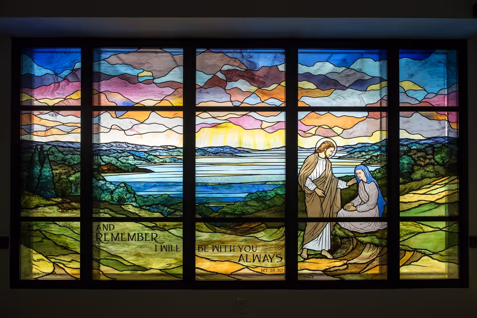 A stained glass window depicting a colorful landscape with hills, a body of water, and a sky with clouds. In the foreground, a figure resembling Jesus is standing and comforting a seated woman dressed in blue and white. The window includes the text: 'AND REMEMBER I WILL BE WITH YOU ALWAYS MT 28:20'.