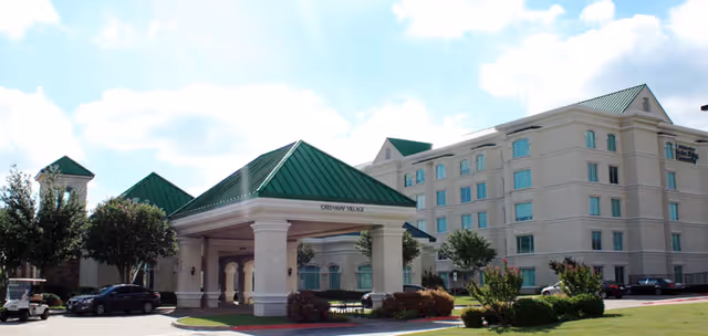 Exterior view of The Courtyard At Christian Care Center showing a large multi-story building with a green roof and a covered entrance labeled Greenway Village. There are trees, shrubs, parked cars, and a clear blue sky with some clouds.