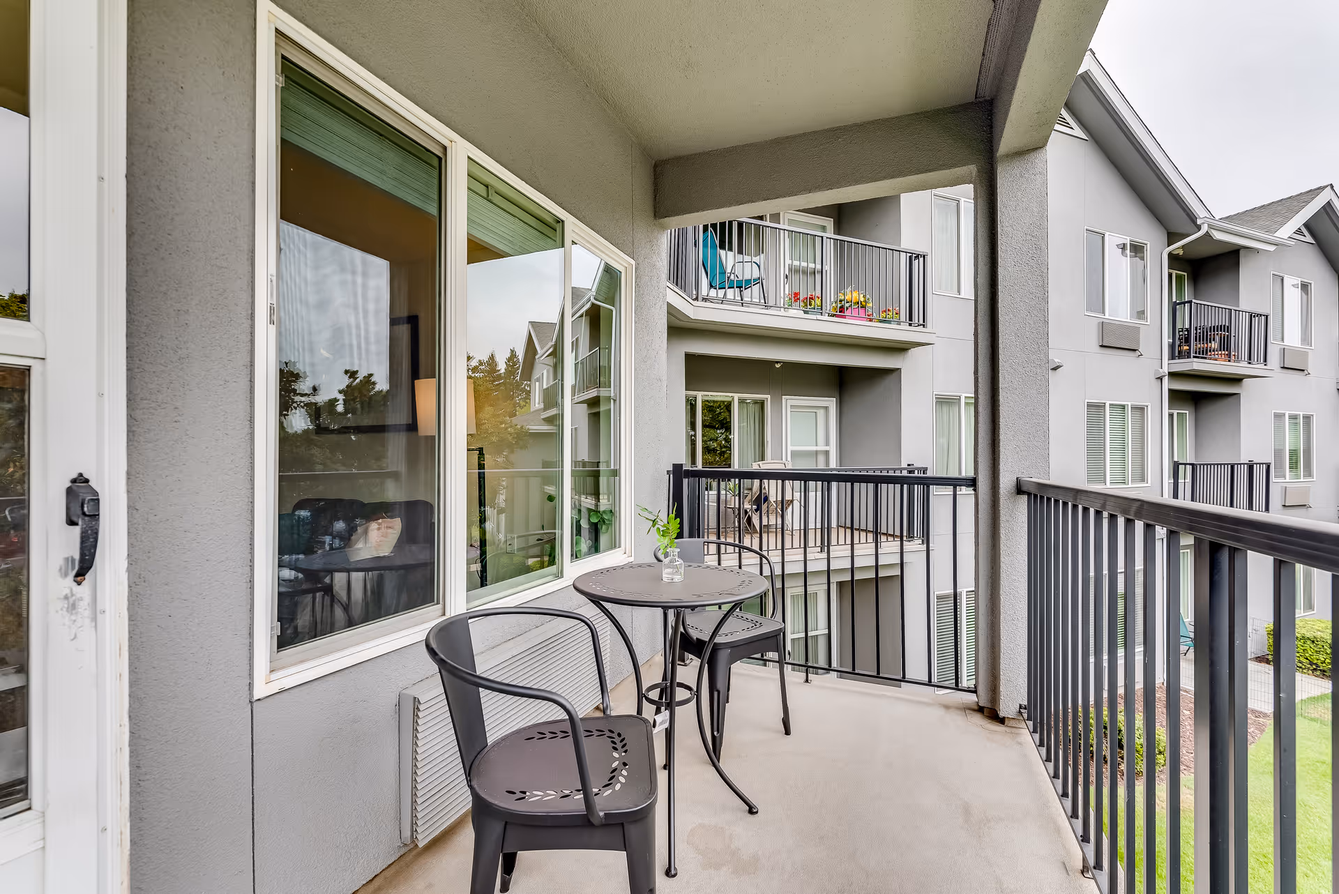 A covered balcony with a small round table and two metal chairs. The balcony overlooks neighboring balconies and a grassy area. The exterior walls and railings are gray, and there is a window and door leading inside the building.