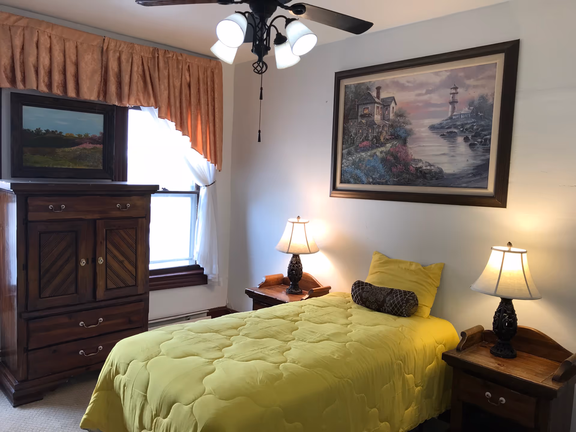 A cozy bedroom with a single bed covered in a yellow quilt and matching pillow. There are two wooden nightstands on either side of the bed, each with a decorative lamp. Above the bed hangs a large framed painting of a coastal scene with a lighthouse. To the left, there is a wooden dresser with a landscape painting on top, and a window with white curtains and a brown valance.