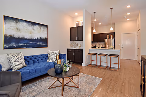 A modern living room and kitchen area featuring a blue tufted sofa with patterned pillows, a round wooden coffee table with decorative vases and plants, a gray armchair, and a large abstract painting on the wall. The kitchen has a breakfast bar with three stools, pendant lighting, stainless steel appliances, and dark cabinetry. The flooring is wood, and the space is well-lit with recessed lighting.