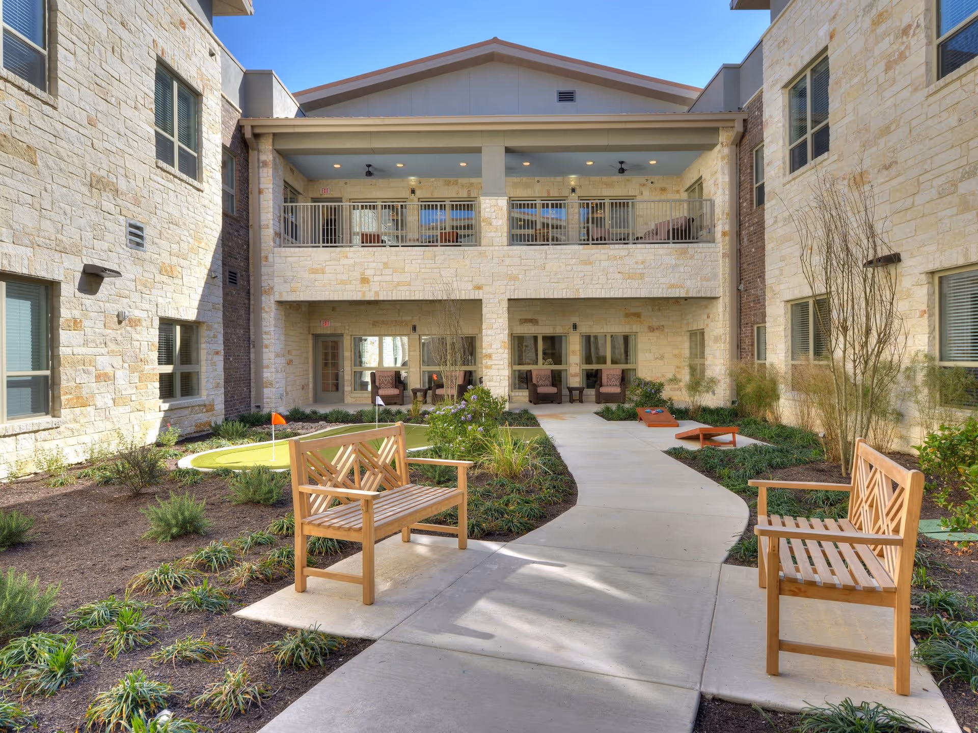 Outdoor courtyard area at Buda Oaks facility featuring a paved walkway flanked by wooden benches, landscaped garden beds with plants, a small putting green with an orange flag, and a cornhole game set. The courtyard is surrounded by a two-story building with stone walls, windows, and a covered balcony with seating on the upper level.