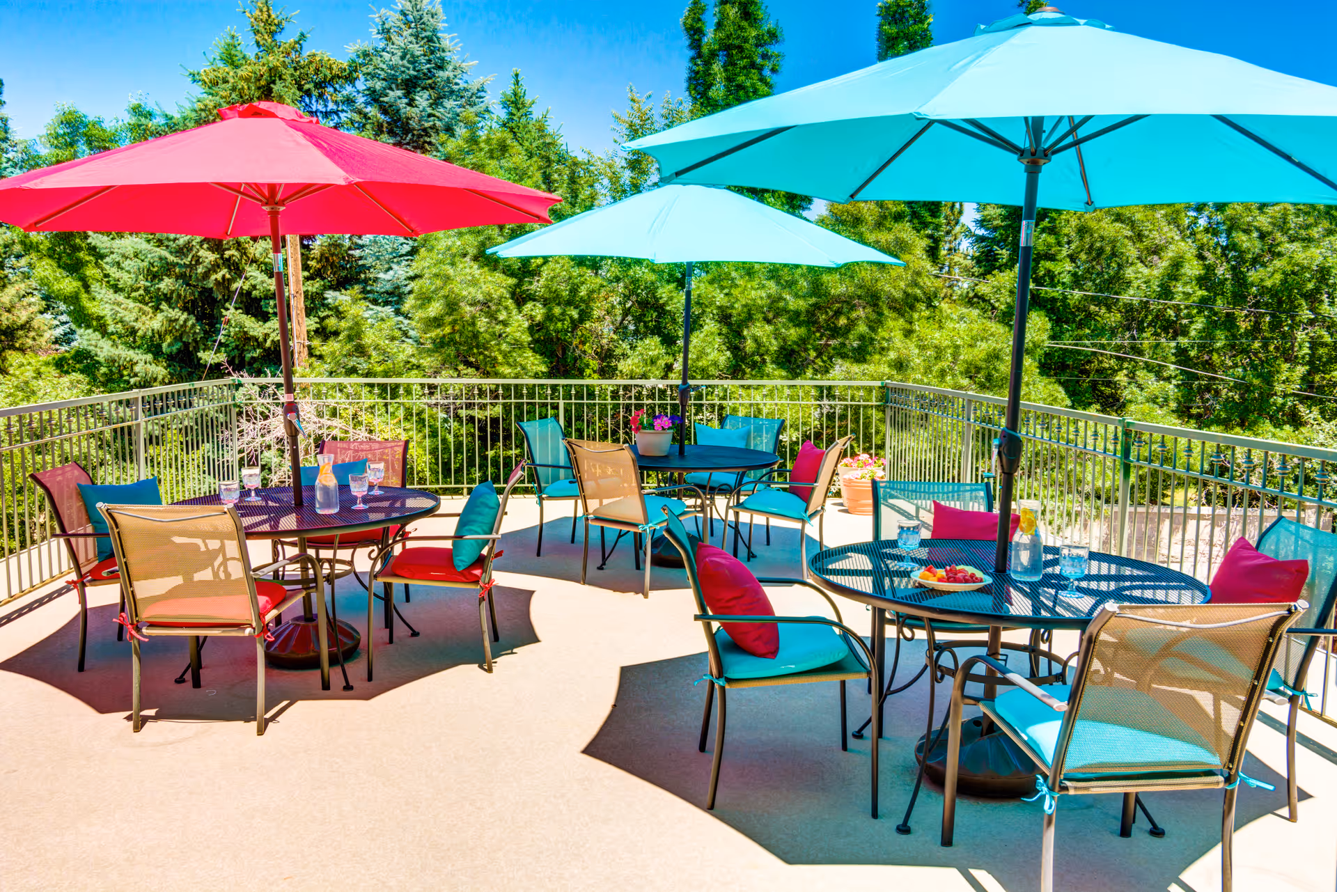 Outdoor patio area with several round metal tables and chairs, each table shaded by large colorful umbrellas in red and blue. The chairs have cushions in teal and red colors. The patio is surrounded by a metal railing and lush green trees in the background under a clear blue sky.