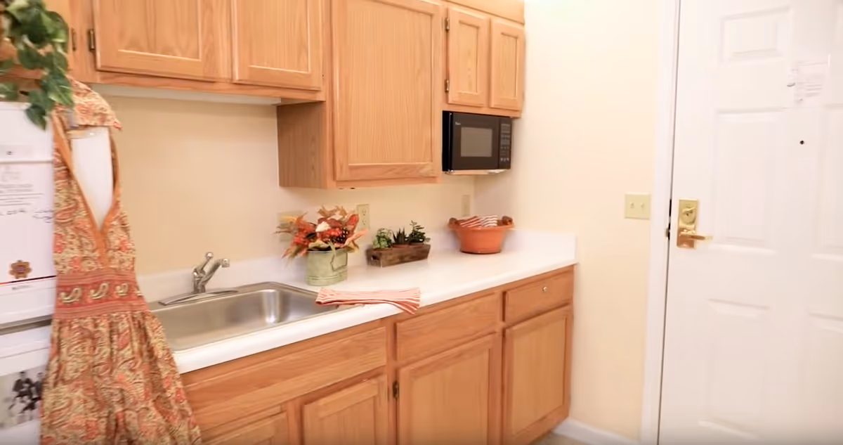 A small kitchen area with wooden cabinets, a stainless steel sink, a microwave mounted above the counter, and a white door to the right. There are decorative plants and a striped cloth on the countertop.