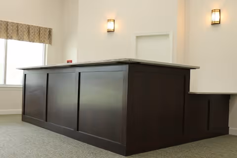 Interior view of a reception or front desk area with a dark wooden counter and a light-colored granite countertop. The room has beige walls with two wall-mounted light fixtures and a window with a patterned valance.