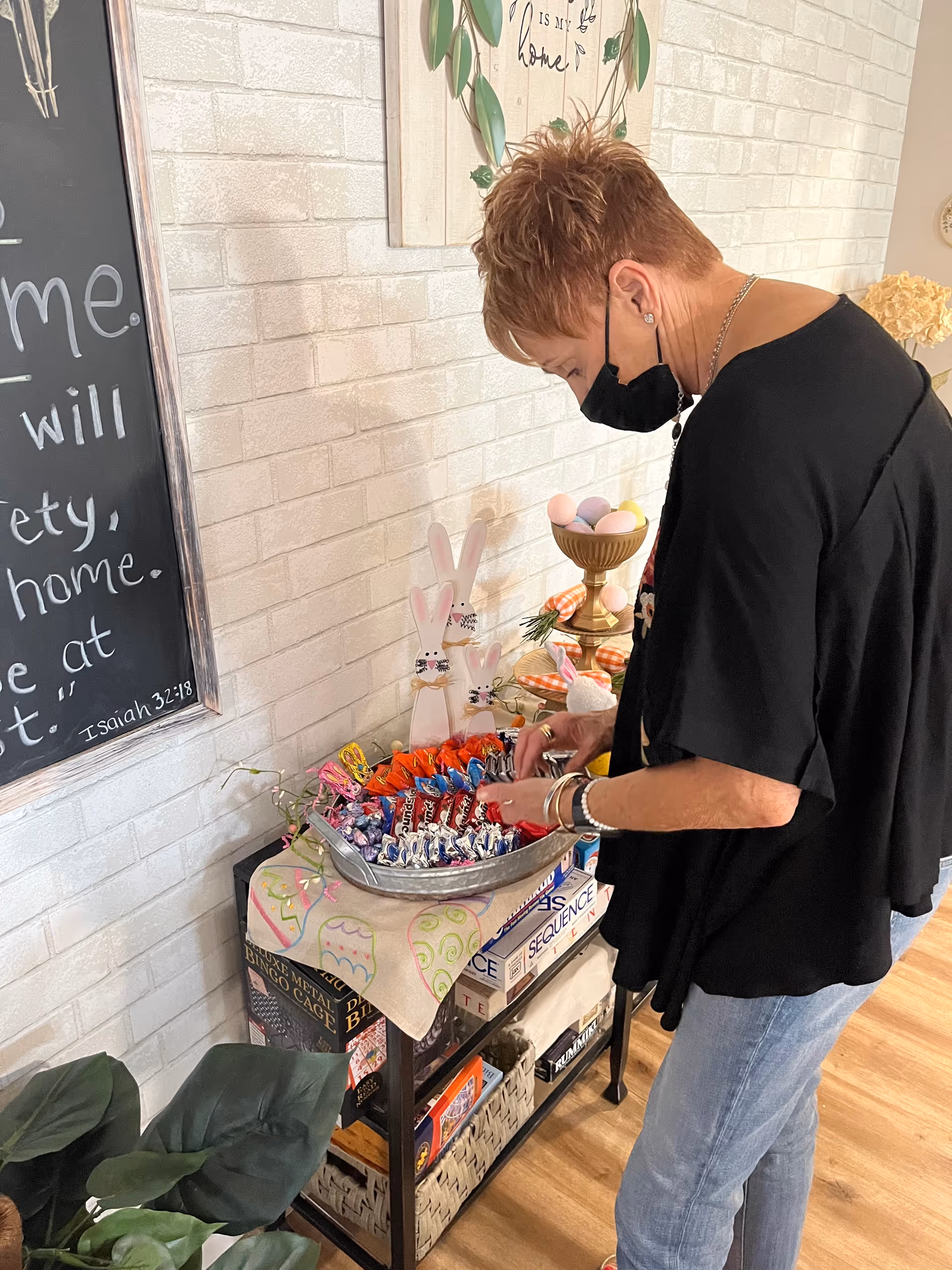 A woman wearing a black mask and black top arranges assorted candy in a metal tray on a small table decorated with Easter-themed items including bunny figures and pastel-colored eggs. The table also holds several board games and is positioned against a white brick wall with a chalkboard and framed artwork.