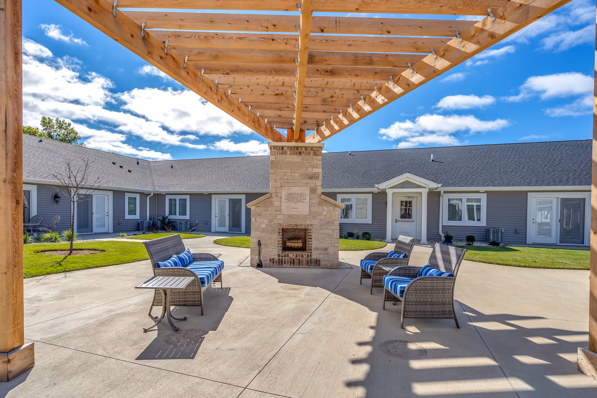 Outdoor courtyard area with a wooden pergola overhead, a stone fireplace in the center, and patio furniture including chairs and a bench with blue and white striped cushions. The courtyard is surrounded by a single-story building with multiple doors and windows, under a bright blue sky with scattered clouds.