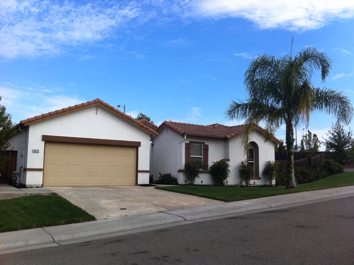 Exterior view of a single-story residential building with a beige garage door, white walls, brown trim, and a red-tiled roof. There is a palm tree and some bushes in the front yard, with a clear blue sky in the background.
