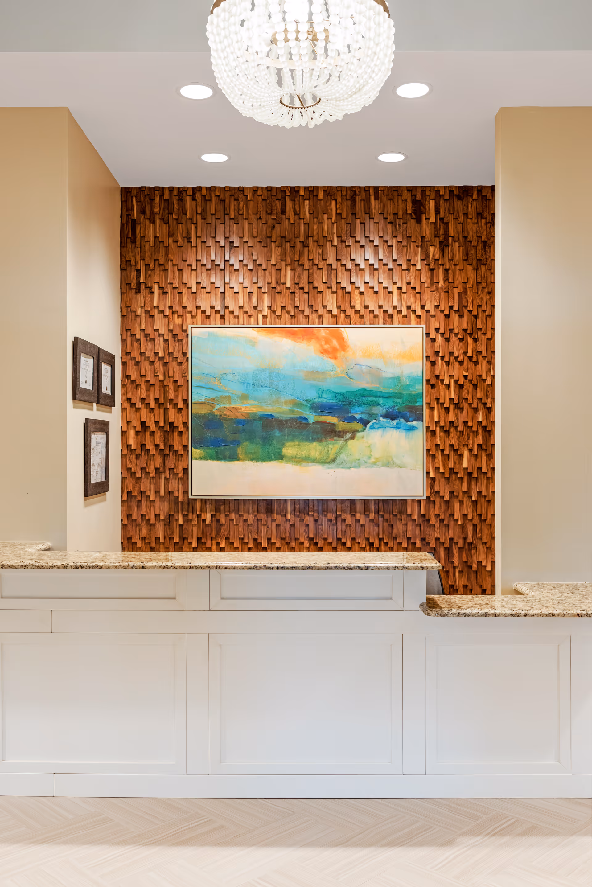 A reception desk in a lobby with a textured wooden accent wall, a colorful painting, and a chandelier overhead.
