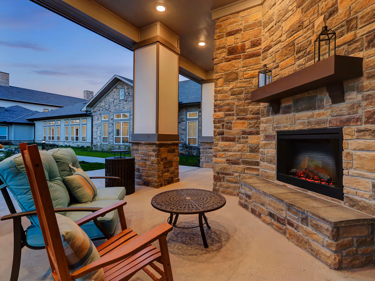 Covered outdoor patio area with cushioned chairs, a small round metal table, and a stone fireplace with a wooden mantel holding two lanterns. The patio overlooks a building with stone and siding exterior walls and multiple windows during dusk.