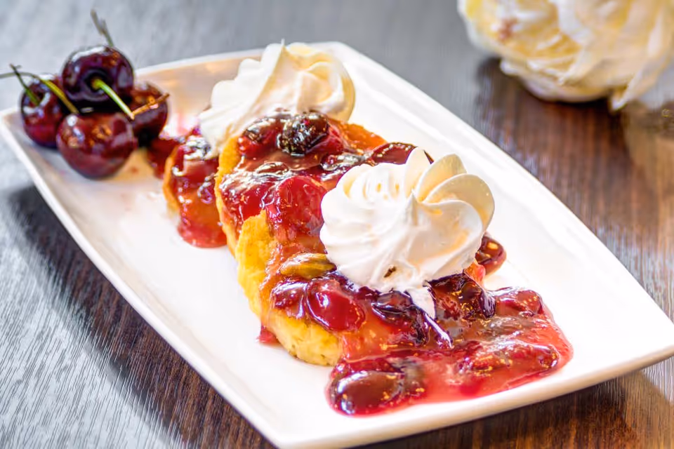 A close-up of a dessert plate featuring three small cakes topped with cherry sauce and whipped cream, with fresh cherries on the side, placed on a wooden table.
