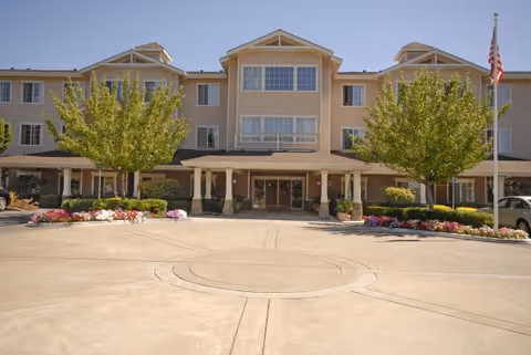 Front exterior view of a three-story senior living facility building with a covered entrance, landscaped flower beds, and trees on either side. An American flag is visible on the right side of the image.
