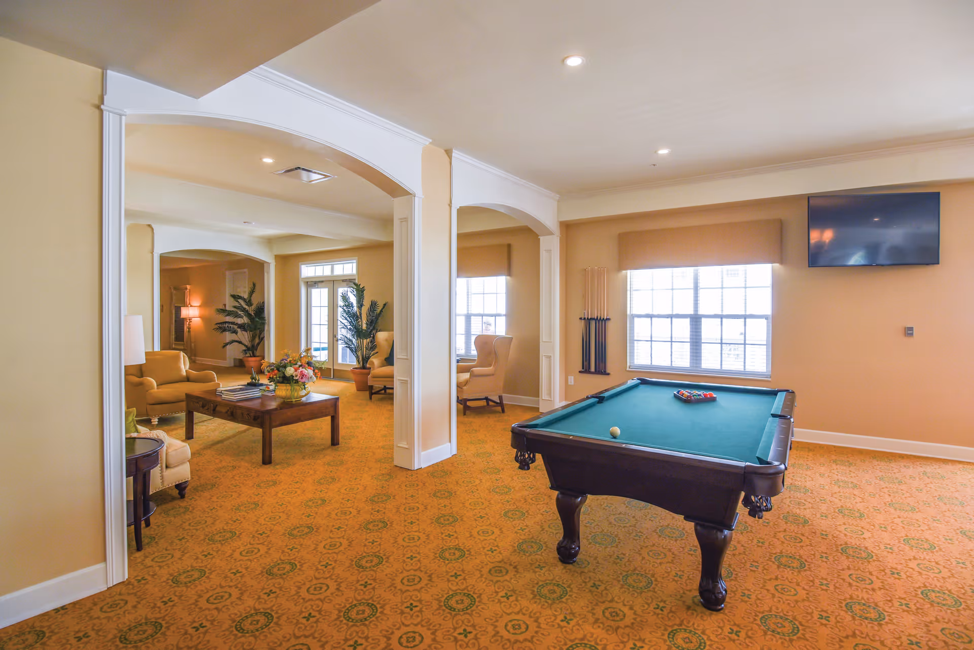 A spacious senior living common area with a pool table in the foreground, a wall-mounted TV, and a seating area with armchairs and a coffee table decorated with a flower arrangement. The room has beige walls, patterned carpet, large windows, and decorative columns.