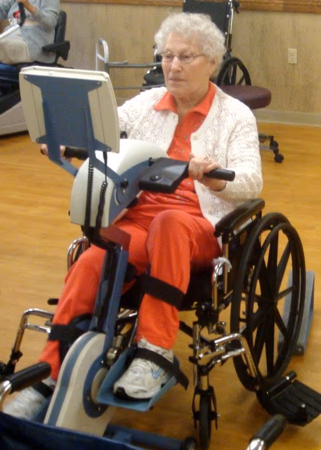 An elderly woman with white curly hair and glasses is seated in a wheelchair using a stationary exercise bike designed for wheelchair users. She is wearing a white knitted cardigan over a red shirt and red pants, with white sneakers. The setting appears to be an indoor facility with wooden flooring and beige walls.