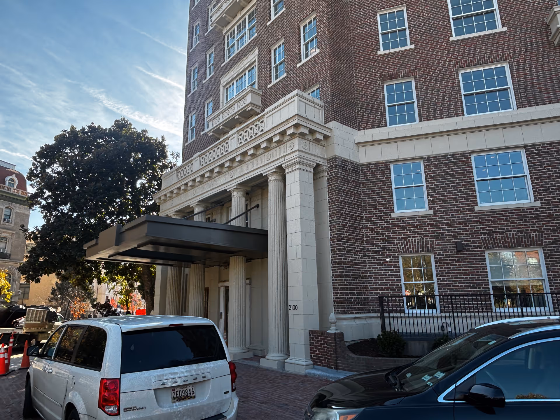 Exterior view of a multi-story brick building with classical architectural details including columns and decorative stonework around the entrance. Two cars are parked in front of the building under a metal canopy. Trees and a clear blue sky are visible in the background.