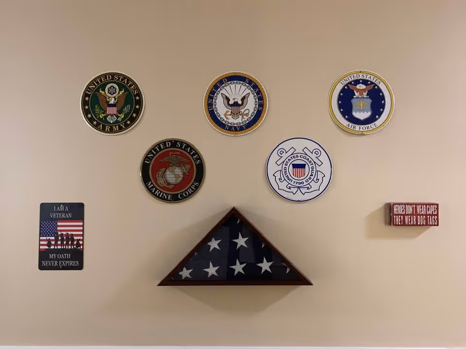 Wall display featuring emblems of the United States Army, Navy, Air Force, Marine Corps, and Coast Guard, with a folded American flag in a triangular wooden case below them. Two plaques are also visible, one with an American flag and the text 'I AM A VETERAN MY OATH NEVER EXPIRES' and another red plaque with the text 'HEROES DON'T WEAR CAPES THEY WEAR DOG TAGS'.
