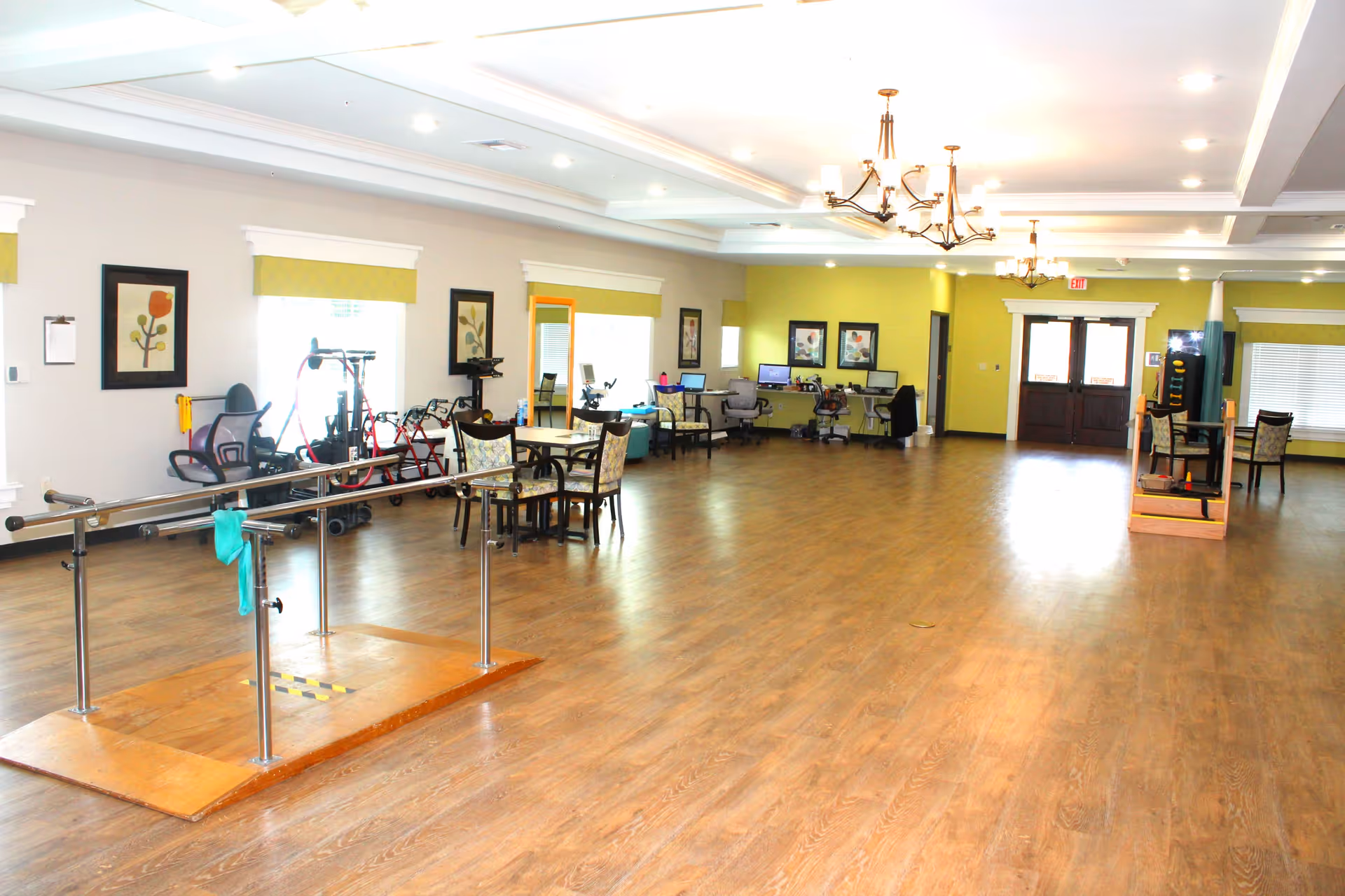 A spacious room with wooden flooring and bright lighting, featuring parallel bars for physical therapy, several chairs and tables, exercise equipment, and computer workstations along the walls. The walls are decorated with framed floral artwork and windows with yellow valances. The room appears to be a rehabilitation or therapy area in a senior living facility.