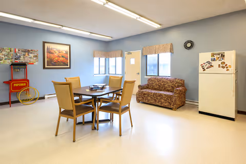 A bright common room with a central table and four chairs, a floral sofa, a refrigerator, and light blue walls with windows.